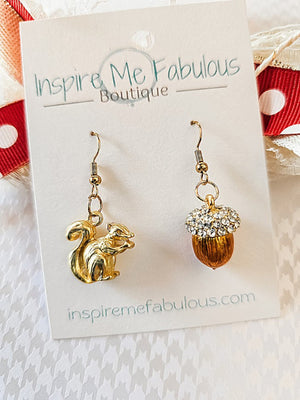 A pair of earrings with one earring featuring a gold-tone squirrel and the other an acorn topped with crystals, displayed on a card with 'Inspire Me Fabulous Boutique' branding.