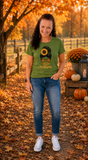 A woman stands outdoors on a leaf-covered path wearing a green graphic T-shirt with a sunflower, turkey, and 'Happy Autumn' design, paired with blue jeans and white sneakers. Pumpkins and autumn trees are visible in the background.