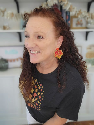 Woman wearing a black t-shirt with a colorful design and orange earrings, sitting in a room with shelves in the background.